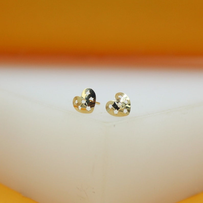 A pair of gold heart-shaped stud earrings featuring intricate star cutouts rests on a white display surface against a warm yellow background.