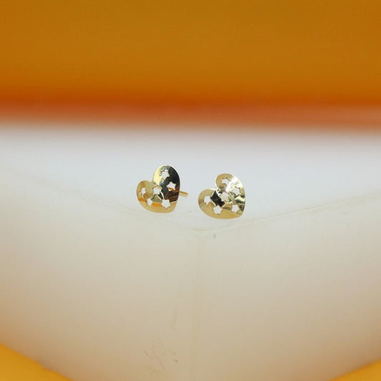 A pair of gold heart-shaped stud earrings featuring intricate star cutouts rests on a white display surface against a warm yellow background.