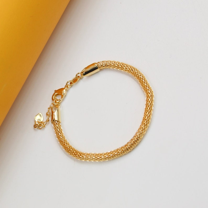 A gold mesh chain bracelet rests on a white surface next to a yellow background.