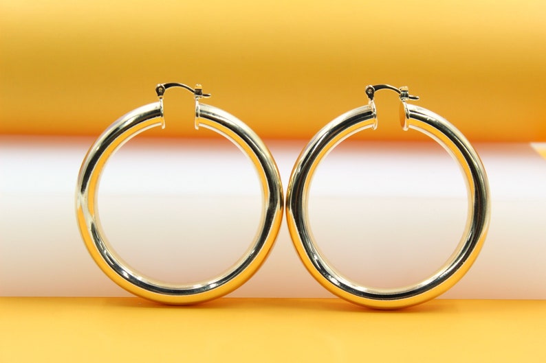 A pair of polished thick gold hoop earrings rests against a minimalist orange and white background.