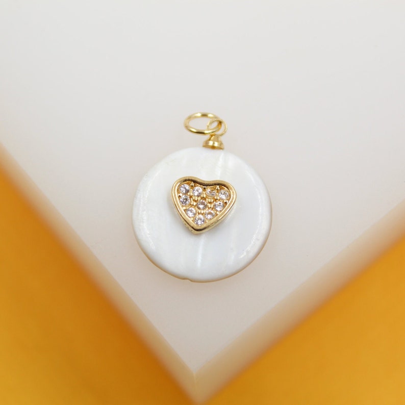 A gold and mother-of-pearl pendant features a sparkling diamond-encrusted heart resting on a white shell circle.