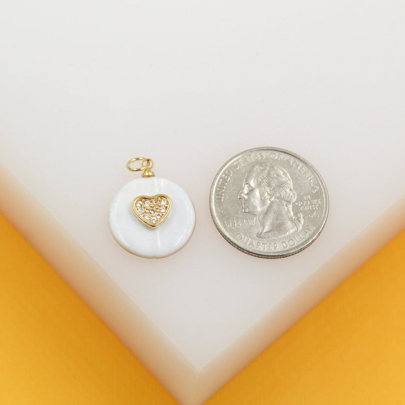 A round white shell pendant with a gold and cubic zirconia heart rests next to a US quarter on a white surface for scale.