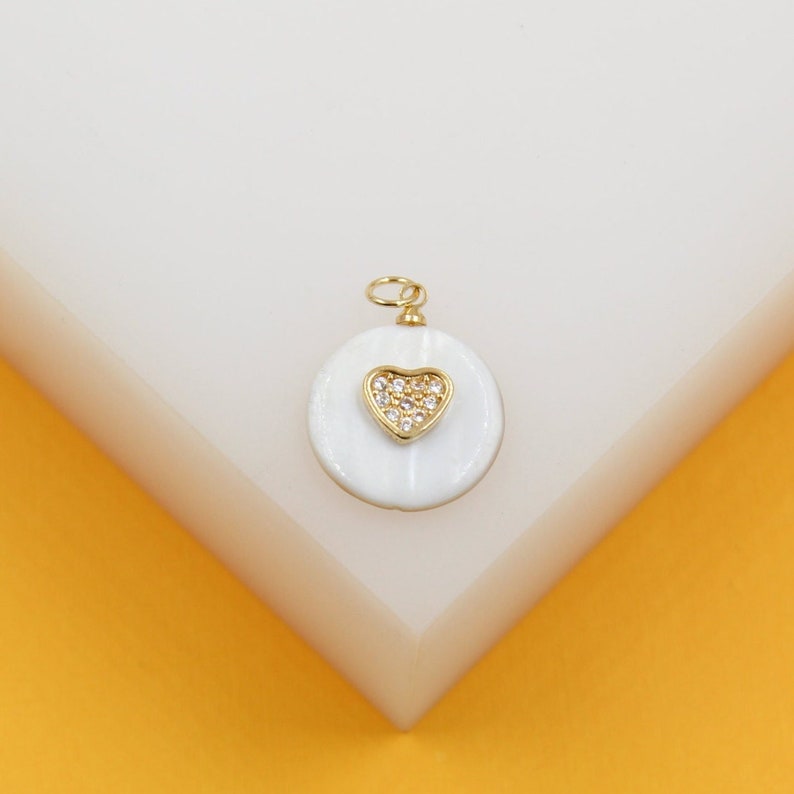A gold-rimmed white pendant featuring a pave diamond heart rests on a white display surface.