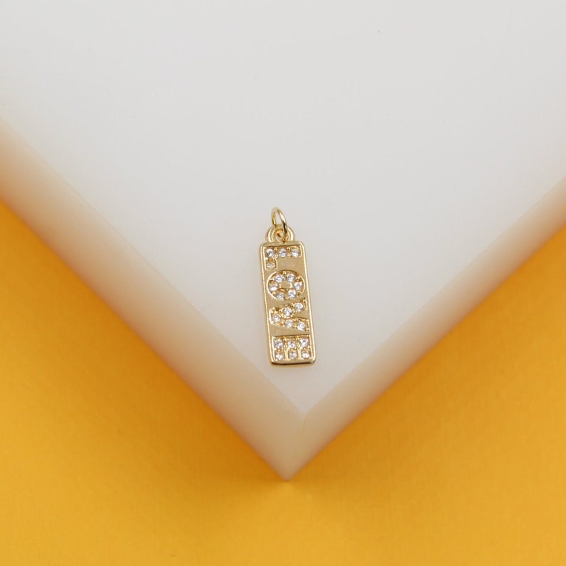 A gold rectangular charm featuring the word "LOVE" spelled out in sparkling cubic zirconia stones rests on a white display surface against a vibrant yellow background.