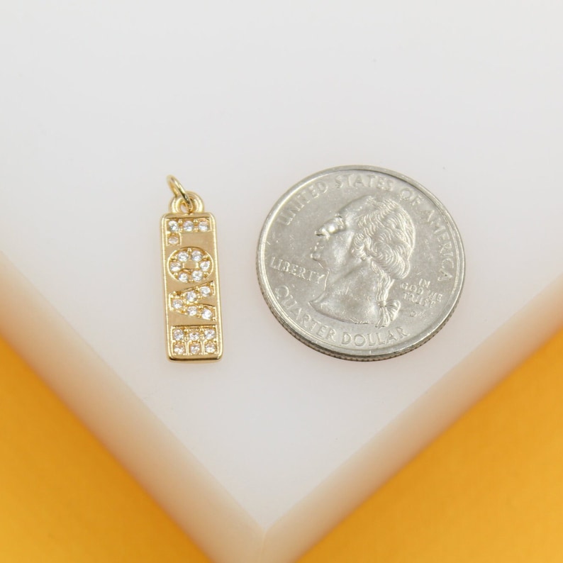 A gold rectangle pendant featuring the word "LOVE" spelled out in sparkling cubic zirconia stones rests next to a US quarter coin for scale.