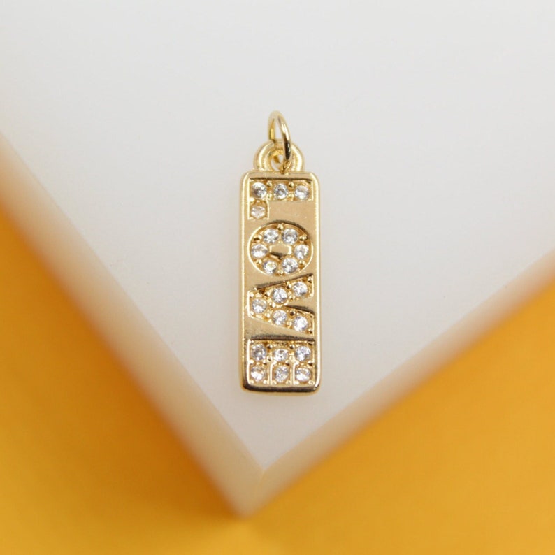 A rectangular gold pendant featuring the word "LOVE" paved with sparkling cubic zirconia crystals rests on a white and yellow background.