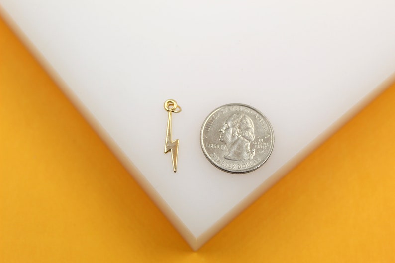 A small gold lightning bolt charm rests next to a US quarter coin on a white display surface to show its scale.