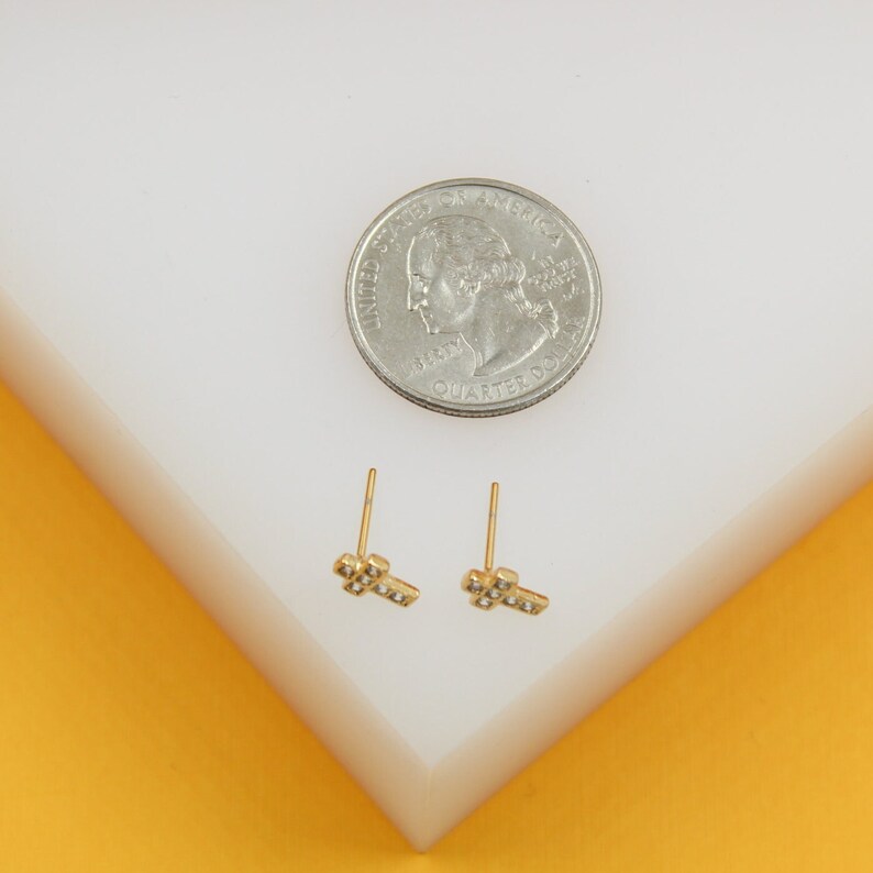 A pair of gold cross stud earrings rests next to a US quarter on a white display stand for size comparison.