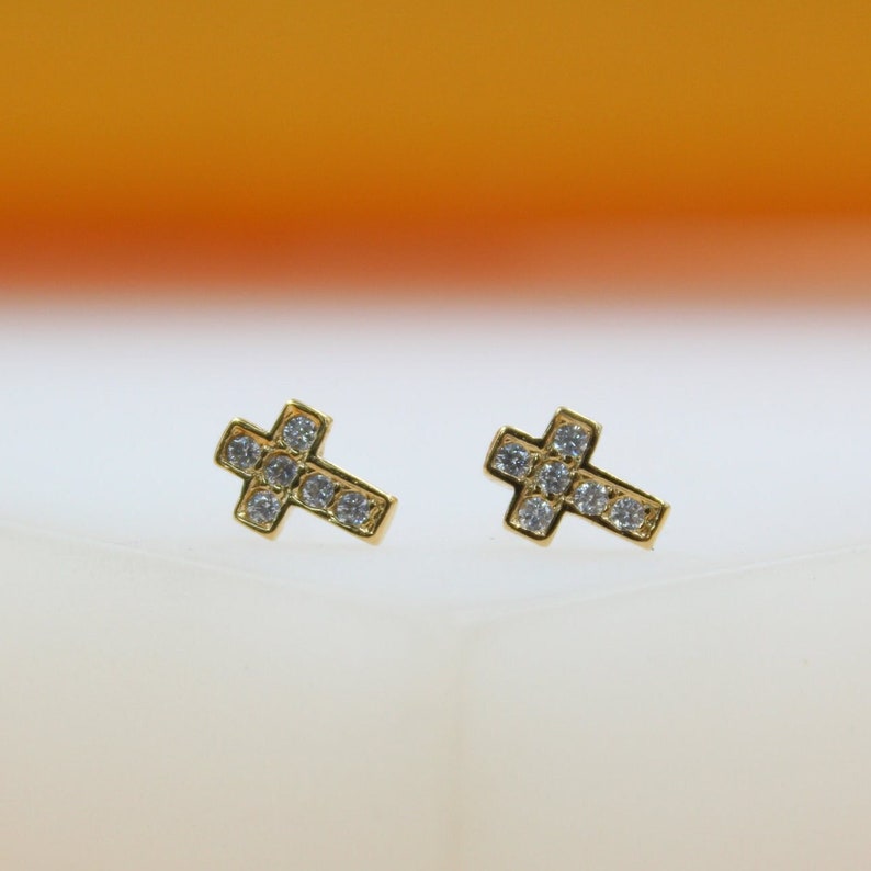 A pair of gold cross stud earrings sparkle with embedded cubic zirconia stones against a soft white and orange background.