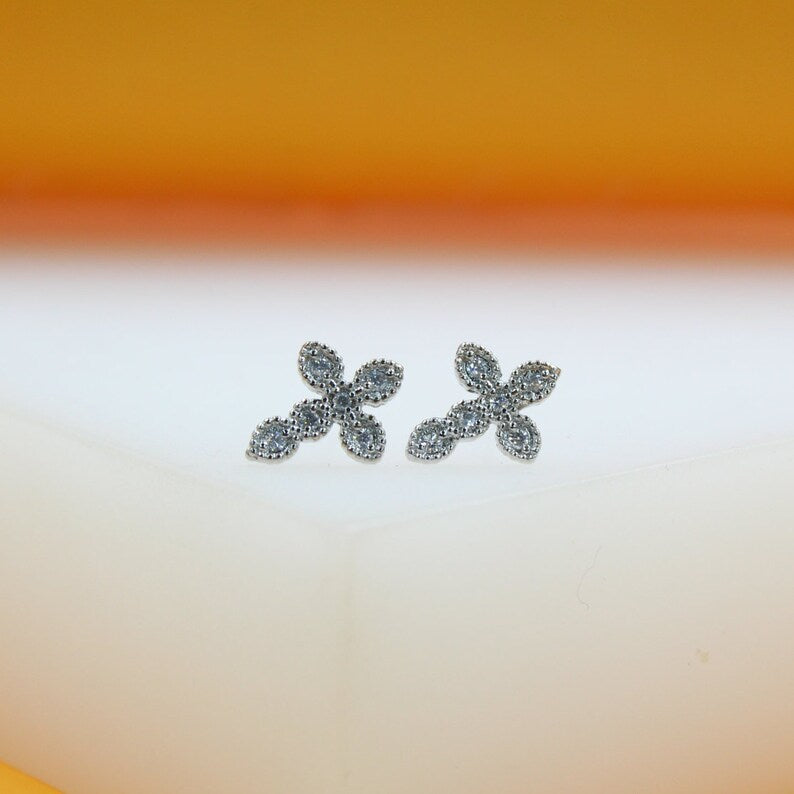 A pair of dainty silver cross stud earrings sparkle with embedded cubic zirconia stones against a soft white and orange background.