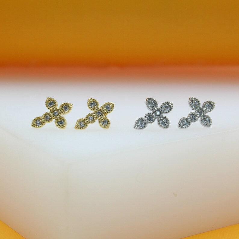 Two pairs of sparkling, flower-shaped cross stud earrings, one in gold and one in silver, rest on a white surface against an orange backdrop.