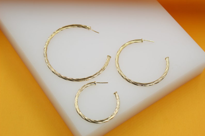 Three twisted gold hoop earrings of varying sizes rest on a white display surface against an orange backdrop.