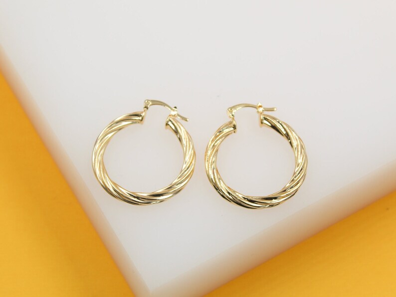 A pair of shiny twisted gold hoop earrings rests on a white and yellow display surface.