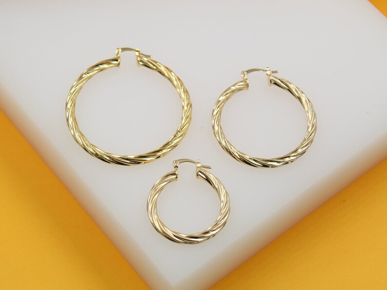 Three gold twisted hoop earrings in graduating sizes rest on a white display stand.