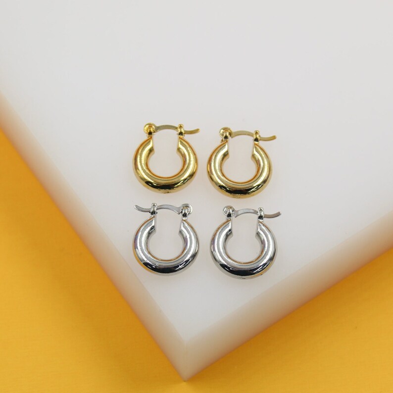 Two pairs of small huggie hoop earrings, one gold and one silver, rest on a white display block against a yellow background.