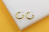 A pair of textured gold hoop earrings rests neatly on a white display stand against a vibrant yellow background.