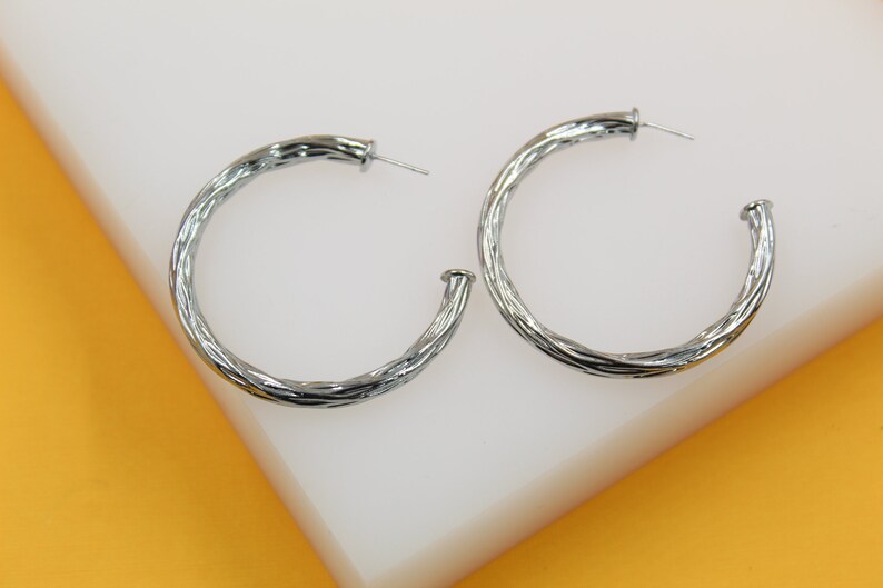Two silver twisted hoop earrings rest on a white display stand against a bright yellow background.