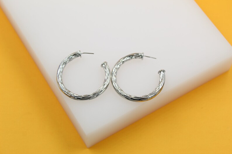 A pair of polished silver twist hoop earrings rests on a white display block against a vibrant yellow background.