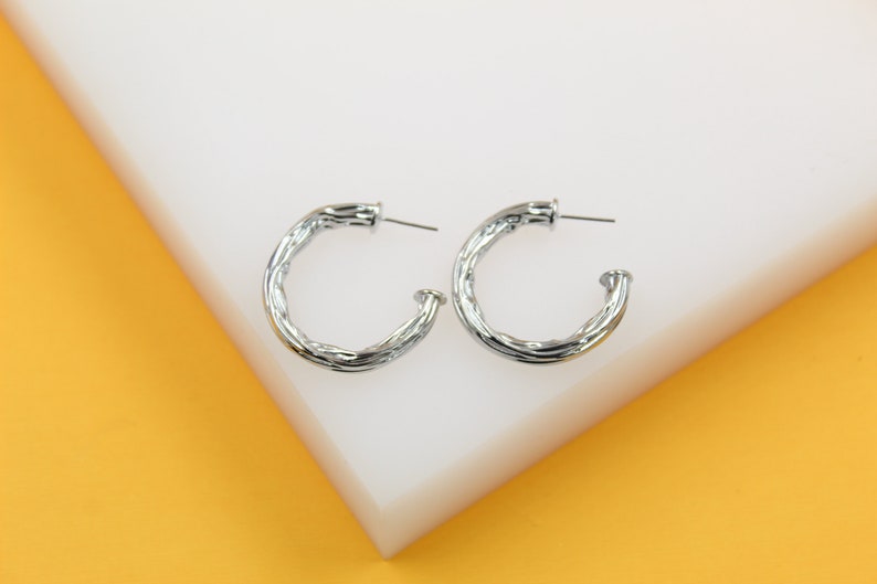A pair of shiny silver twisted hoop earrings rest neatly on a white and yellow display block.