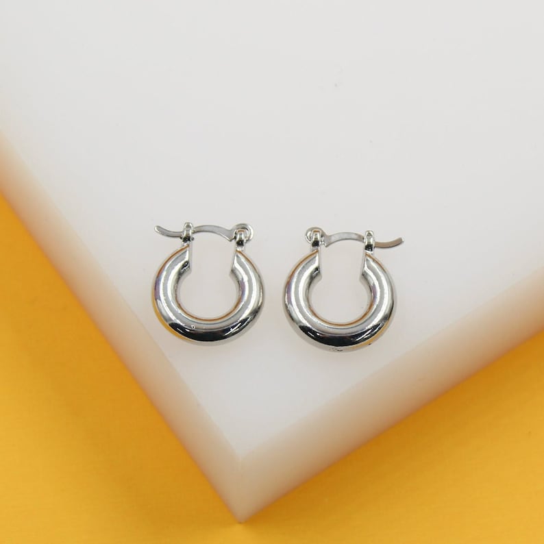 A pair of polished silver hoop earrings rests neatly on a white display stand against a warm yellow background.