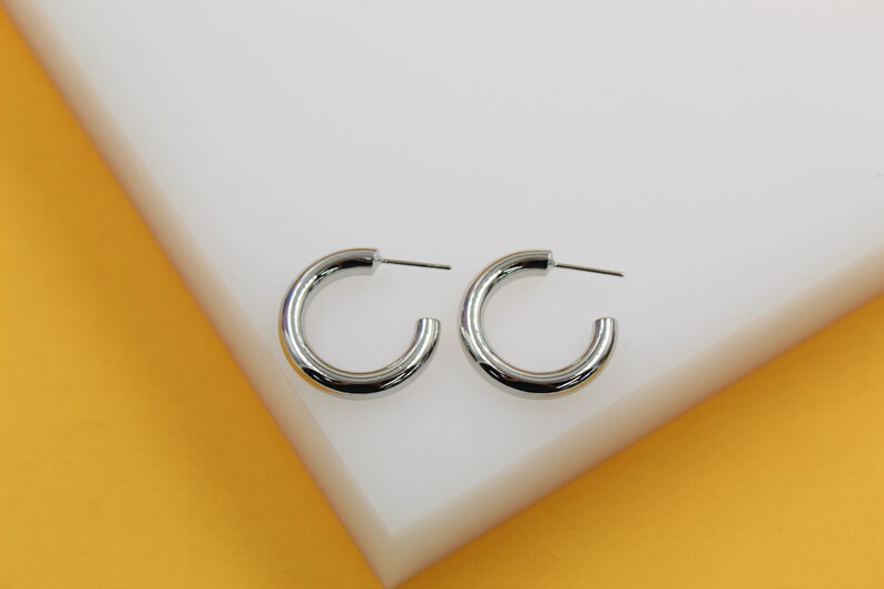 A pair of shiny silver hoop earrings rests neatly on a white display surface against a vibrant yellow background.