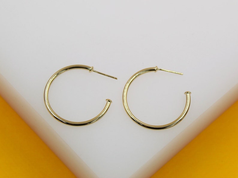 A pair of polished gold hoop earrings rests neatly on a clean white surface against an orange backdrop.