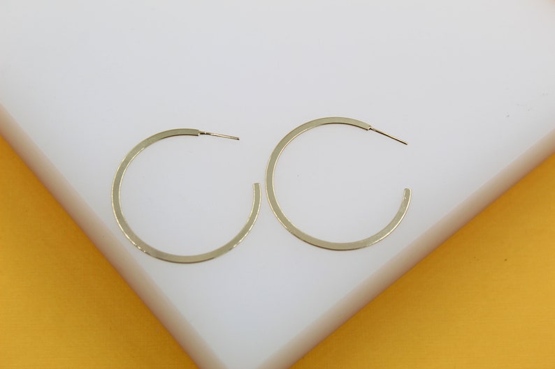 A pair of minimalist gold hoop earrings rests neatly on a clean white display surface against a vibrant yellow background.