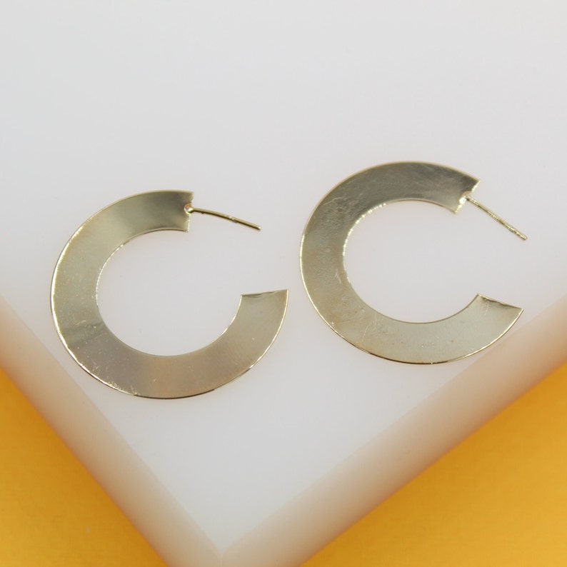 A pair of wide, flat gold hoop earrings rests on a white and yellow surface.