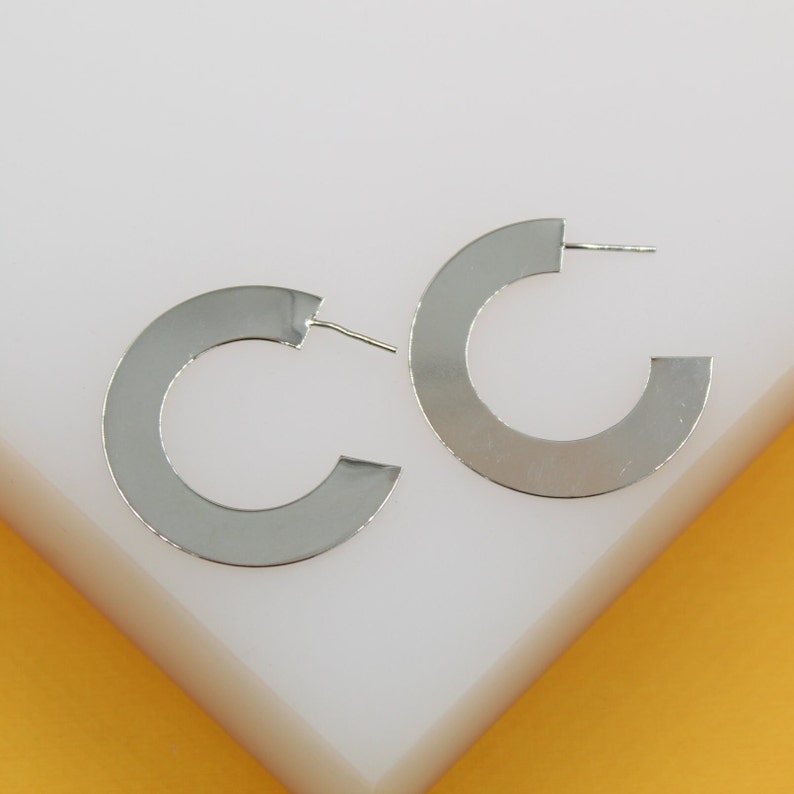A pair of sleek silver wide flat hoop earrings rests on a white and yellow display block.