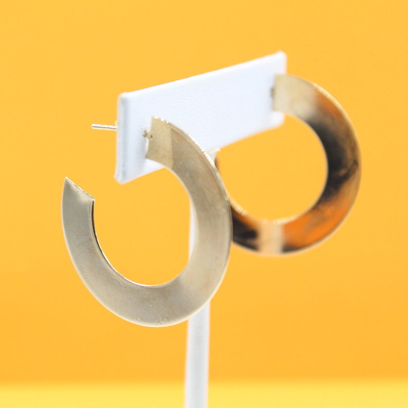 A pair of wide, flat gold hoop earrings rests on a white display stand against a bright yellow background.