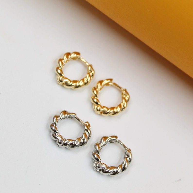 Two pairs of twisted gold and silver hoop earrings rest on a white surface against a warm yellow backdrop.