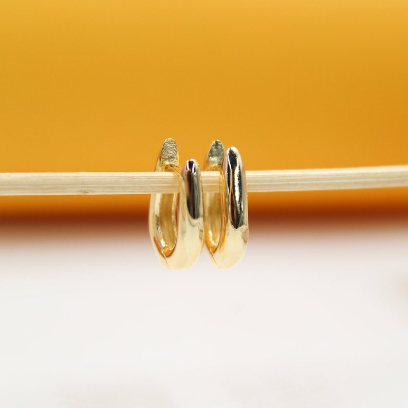 A pair of polished gold hoop earrings hangs delicately on a wooden stick against a warm yellow and white backdrop.