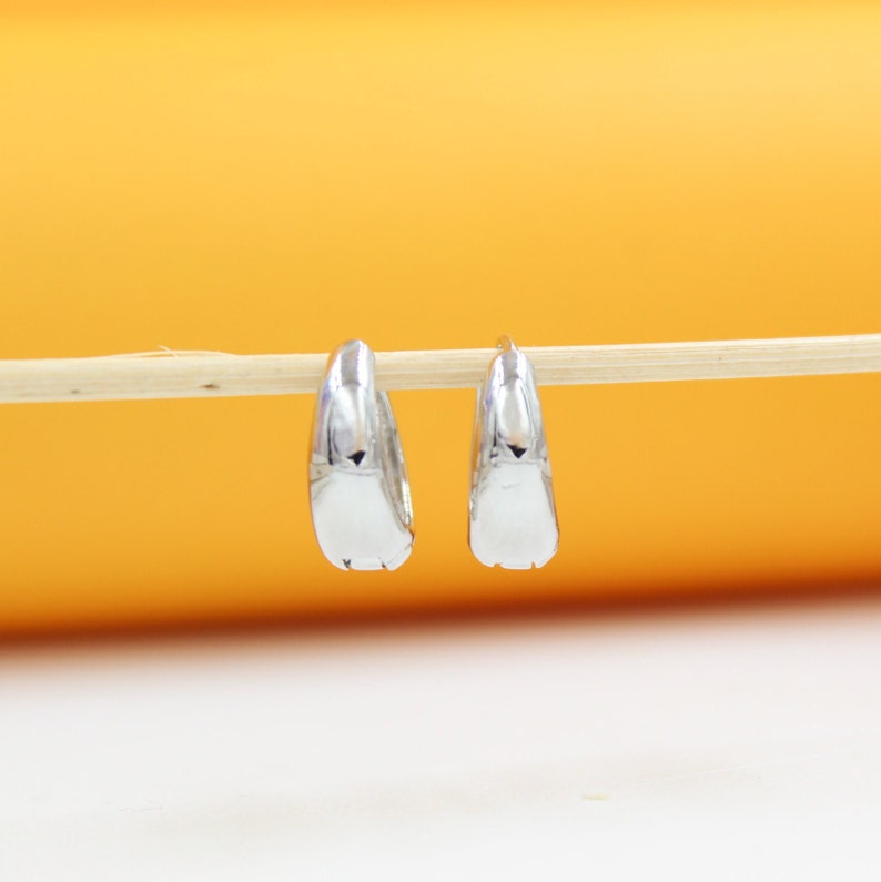 A pair of polished silver hoop earrings hangs delicately from a wooden stick against a warm orange background.