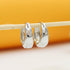 A pair of polished silver huggie hoop earrings hangs neatly from a thin wooden stick against a warm yellow and white background.