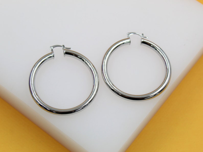 A pair of sleek silver hoop earrings rests on a white display surface against a warm yellow background.