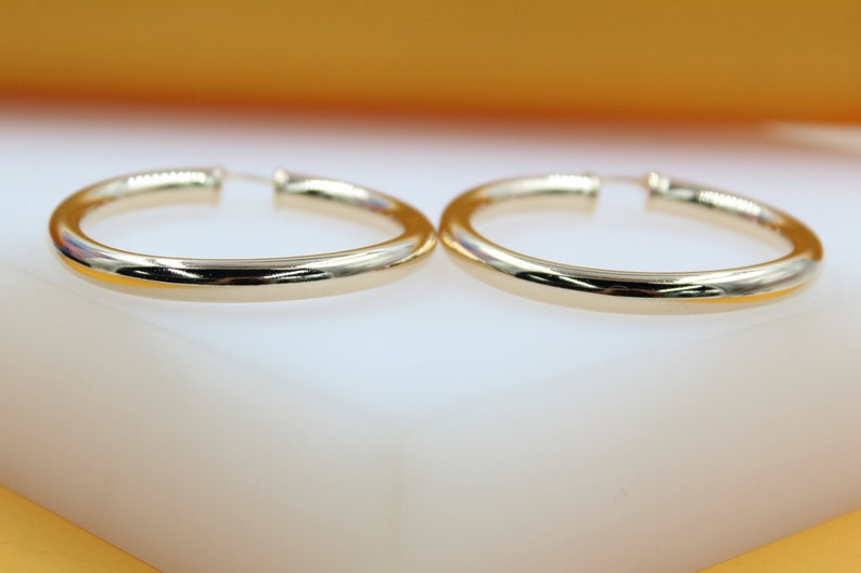 A pair of polished gold hoop earrings rests neatly on a white surface against a warm yellow backdrop.