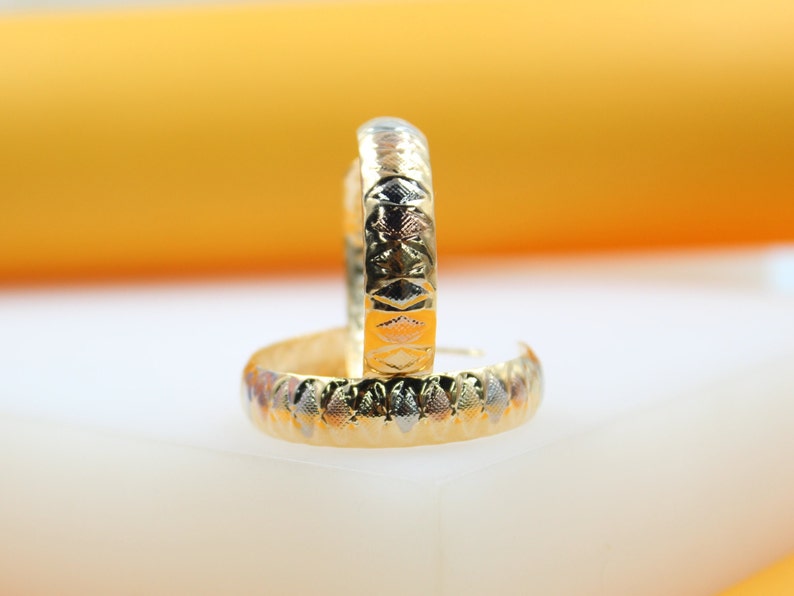 A pair of gold-plated textured hoop earrings rest elegantly on a white surface against a warm yellow background.