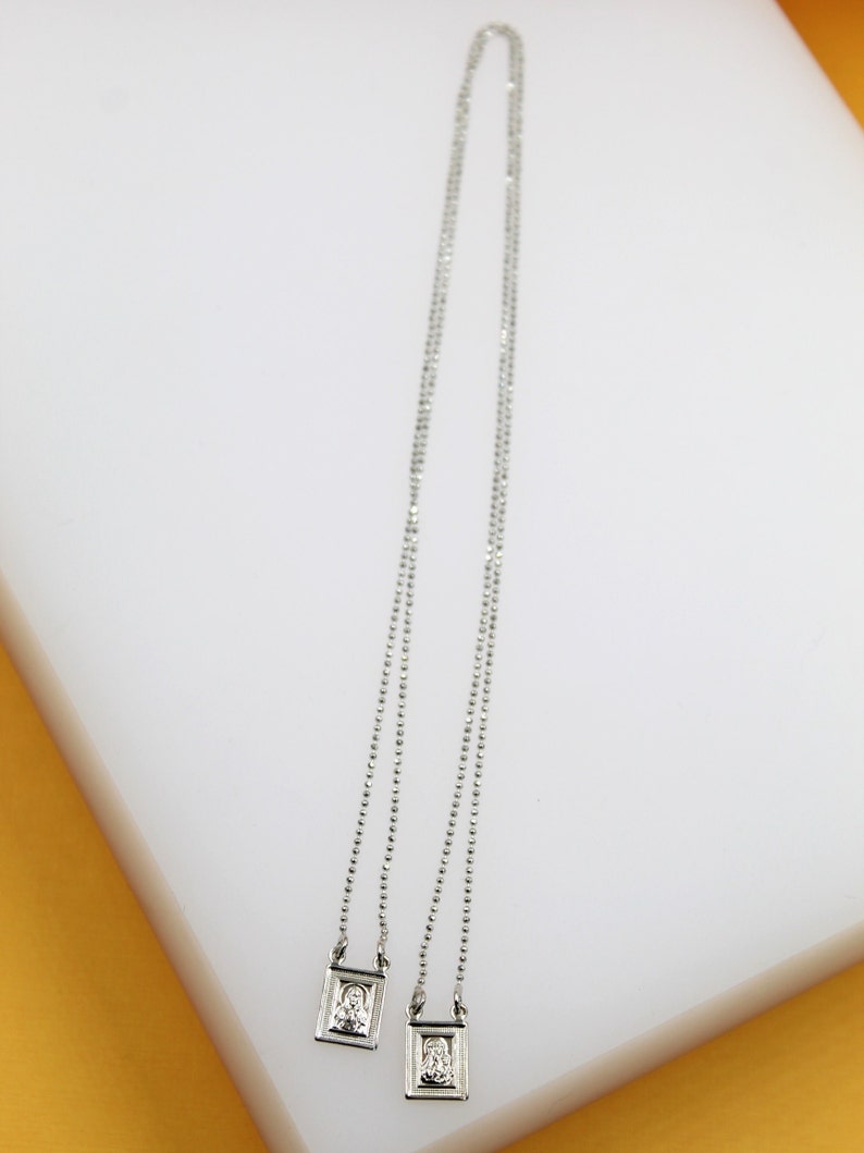 A delicate silver scapular necklace with two small rectangular pendants rests on a white display surface.