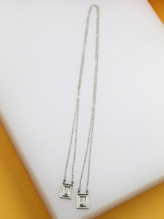 A delicate silver scapular necklace with two small rectangular pendants rests on a white display surface.