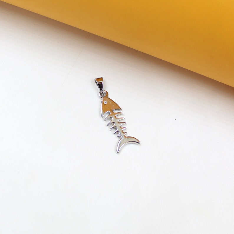 A shiny silver fish skeleton pendant rests on a clean white and yellow background.