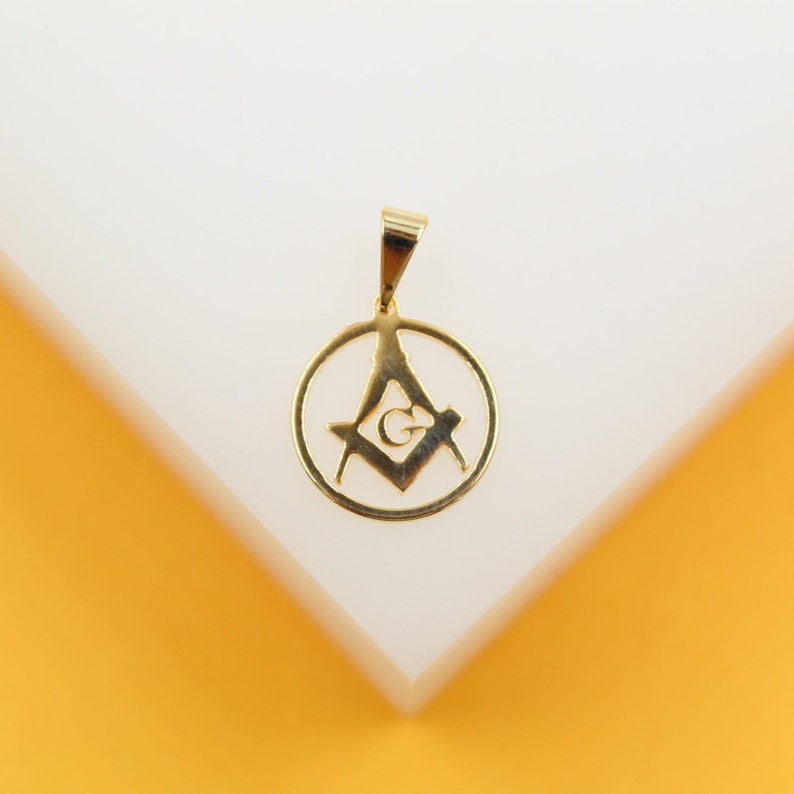 A polished gold Masonic pendant featuring the square and compasses symbol with the letter G rests on a white display surface.