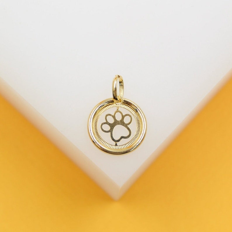 A gold paw print charm pendant rests on a clean white display surface against a vibrant yellow background.