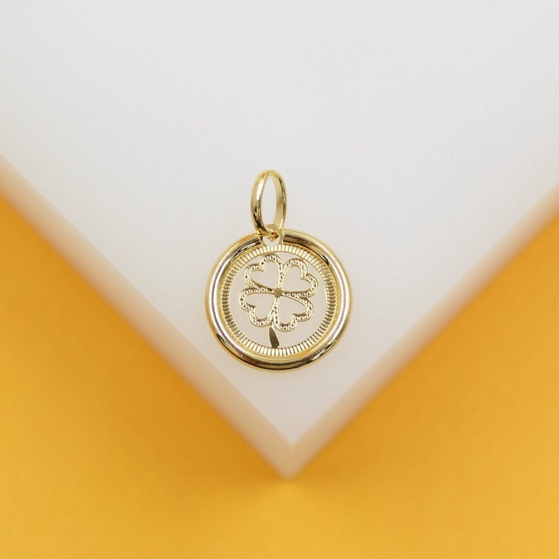 A delicate gold pendant featuring a four-leaf clover cutout rests on a white display surface against a warm yellow background.