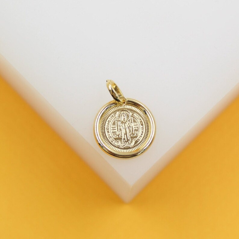 A polished gold Saint Benedict medal pendant rests on a clean white and yellow display surface.