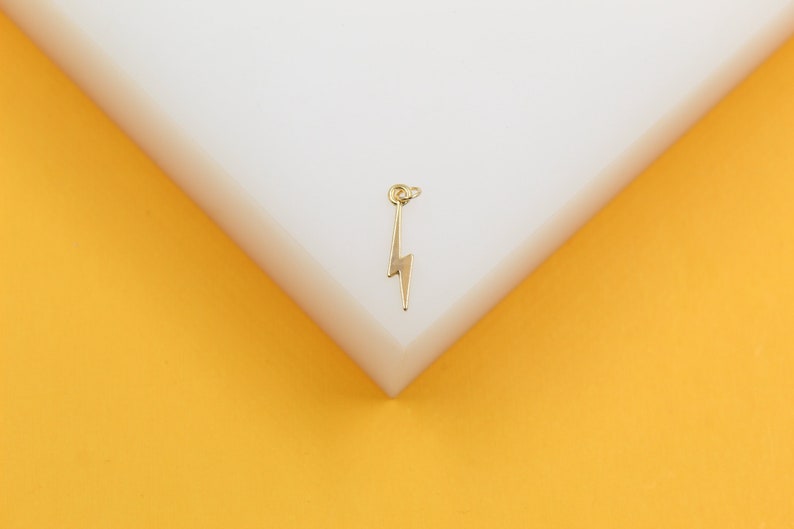 A delicate gold lightning bolt charm rests on a white display surface against a vibrant yellow background.