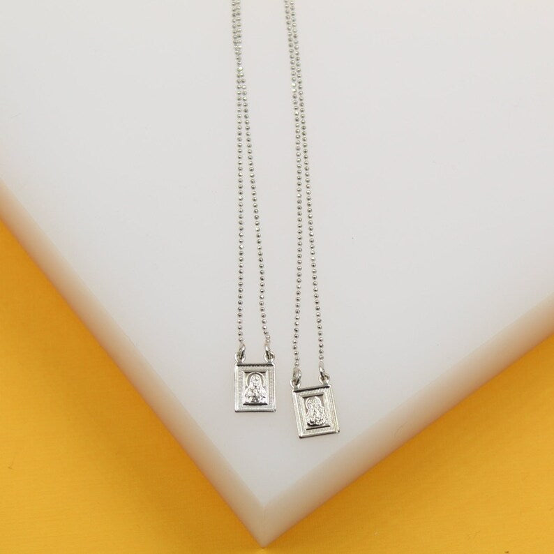Two delicate silver scapular necklaces with rectangular pendants lie side by side on a white and yellow display stand.
