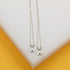 Two delicate silver scapular necklaces with rectangular pendants lie side by side on a white and yellow display stand.