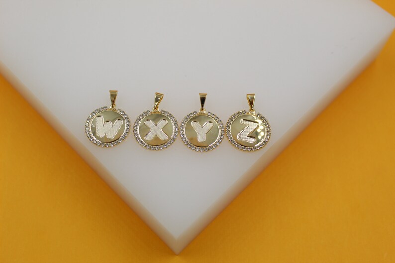 Four gold round pendant charms featuring the letters W, X, Y, and Z surrounded by rhinestones lie on a white display stand.