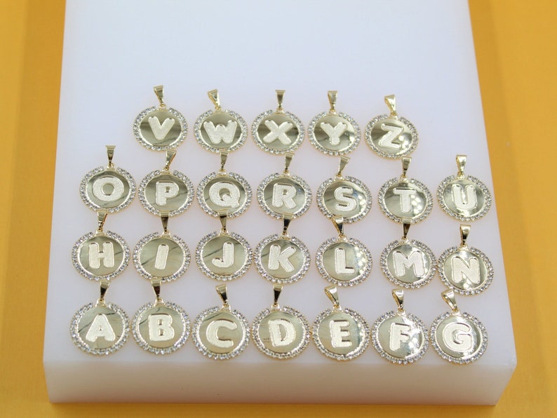 A collection of gold initial letter pendants, encrusted with rhinestones, are arranged in alphabetical order on a white display block.