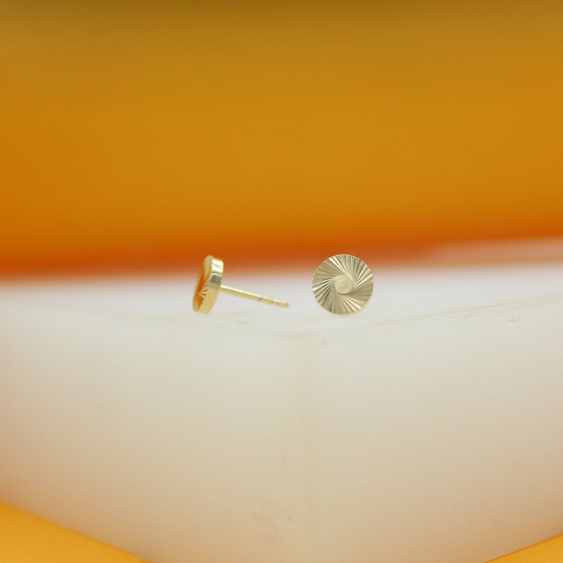 A pair of round, textured gold stud earrings rests on a white surface against an orange background.